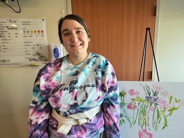 Rose Principe, wearing a tie-dye top, smiles next to painting of flowers she did in therapy.