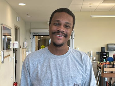 Demarius Branson smiling while standing in therapy gym and wearing a gray shirt.