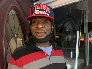 Willis Rogers standing in a doorway wearing a hat that says St. Louis, Missouri and striped shirt.