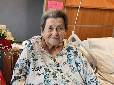 Suzanne Gregg sits and smiles at the camera from her hospital room sitting in a chair next to the bed.